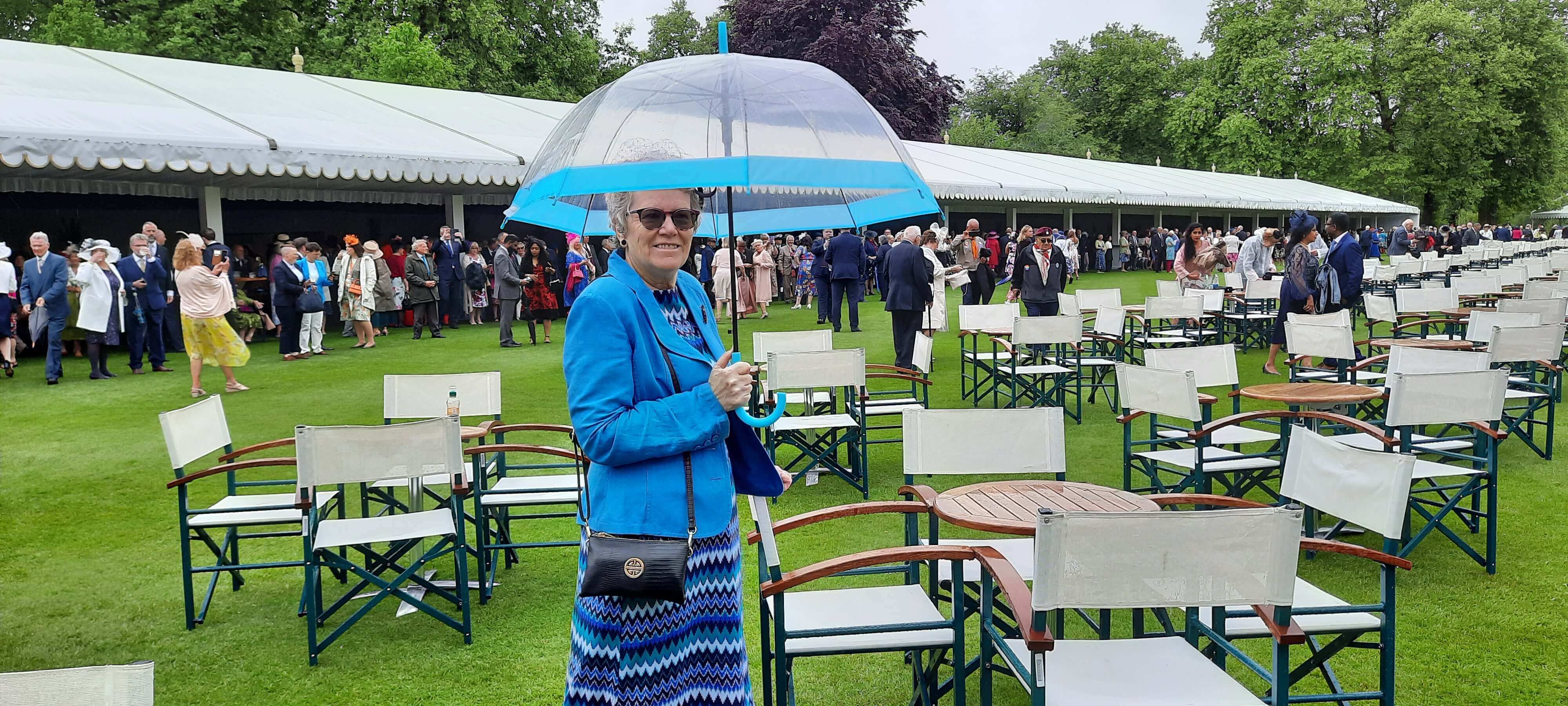 An ROS volunteer standing in front of a marquee on the lawns of Buckingham Palace with a blue umbrella