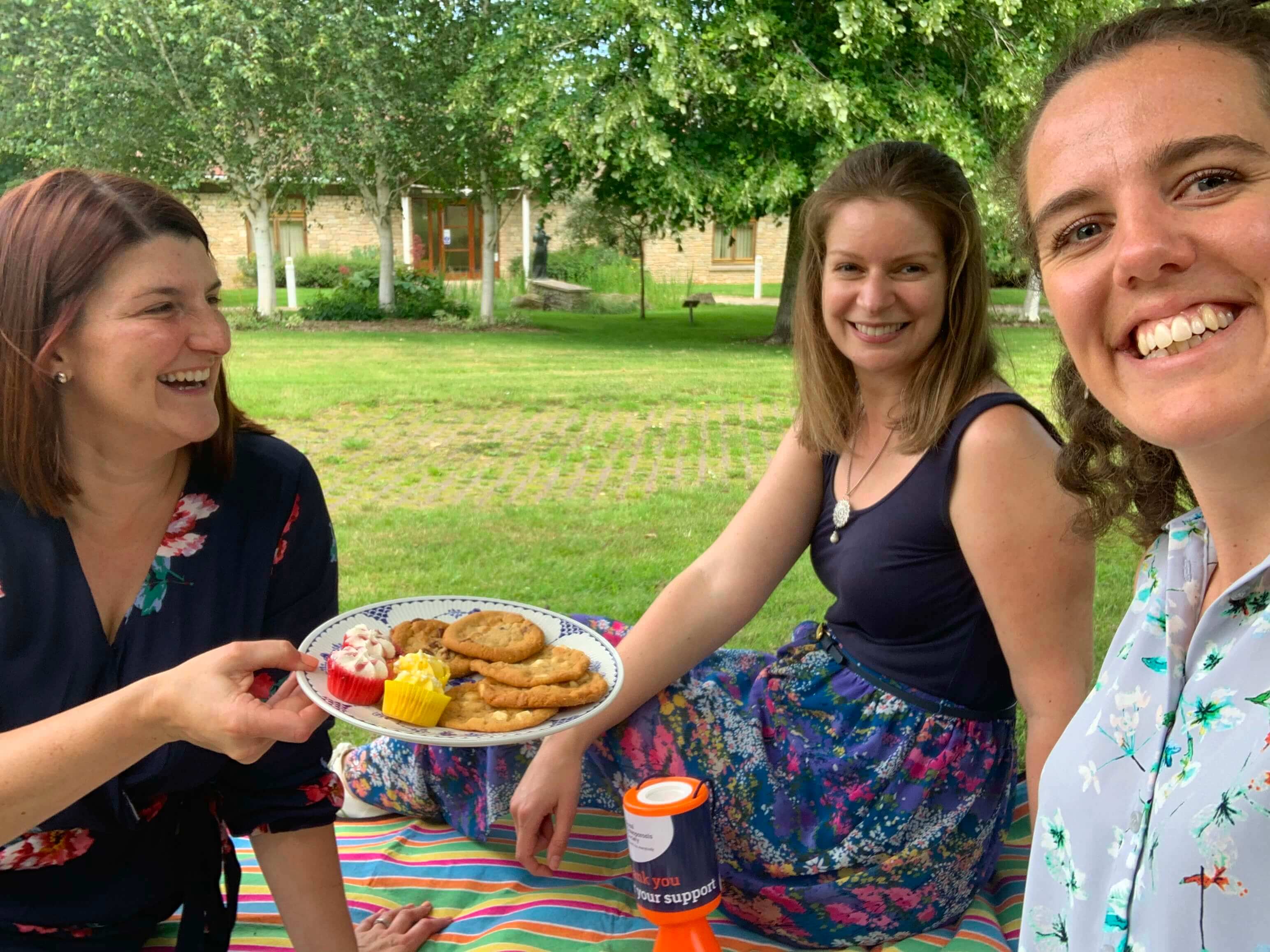 Three people sat outside on the grass with a plate of cakes, smiling