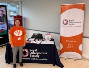 A volunteer in an orange ROS t-shirt stands next to a banner and table of leaflets